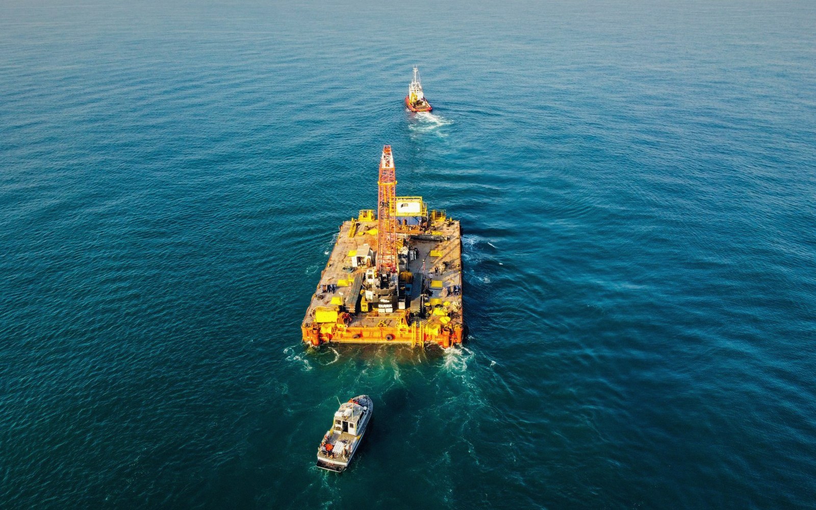 Aerial view of a barge with a drilling rig anchored at sea, flanked by two boats
