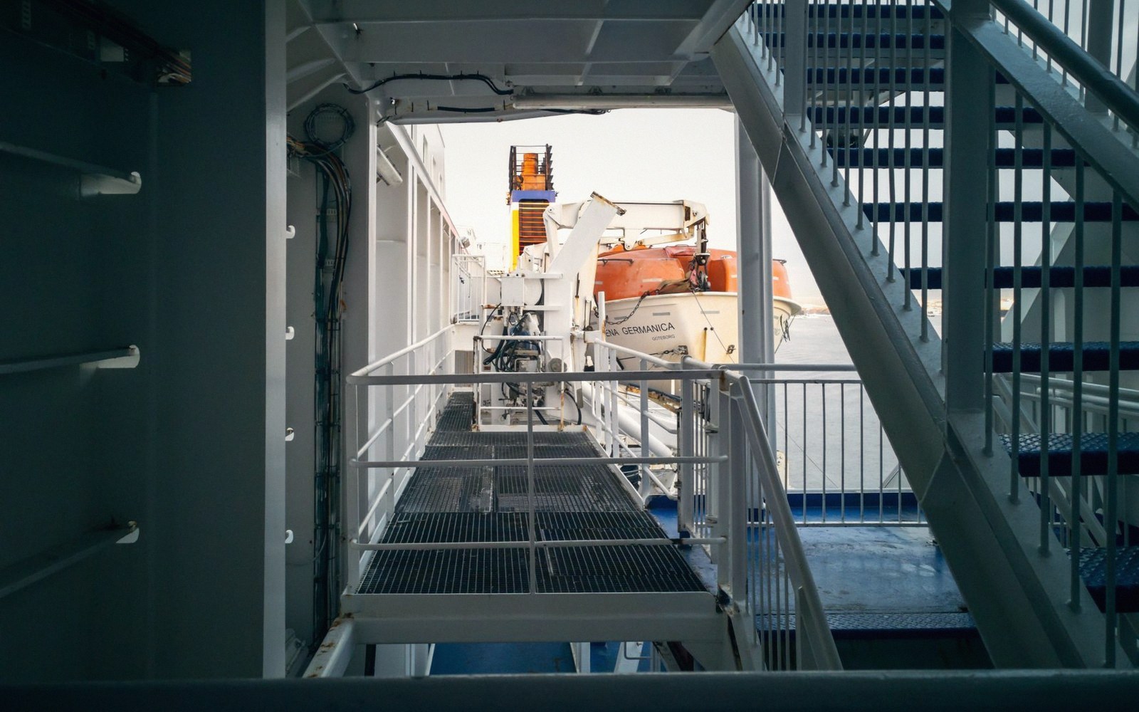 Metal walkway and staircase aboard a ferry, with an enclosed lifeboat visible outside