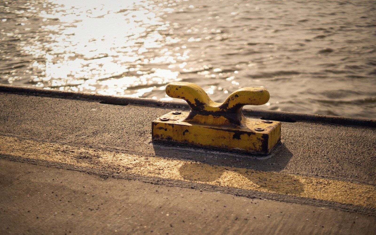 A weathered yellow bollard sits on a concrete dock beside sunlit rippling water