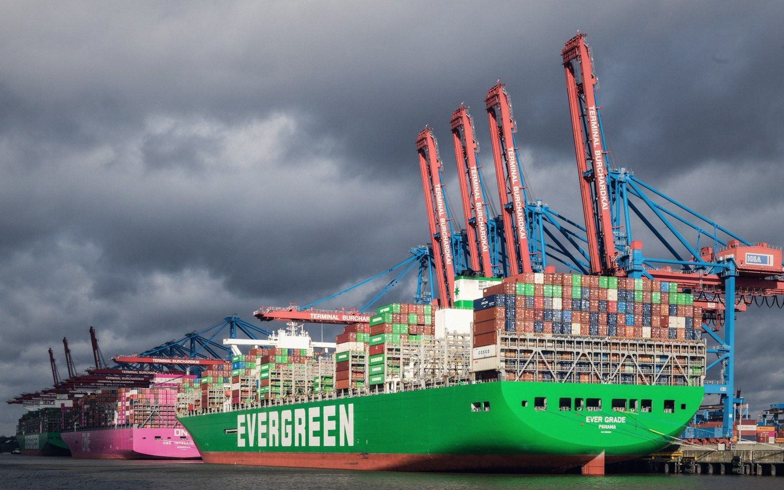 An Evergreen container ship loaded with cargo docked beneath tall cranes under a cloudy sky