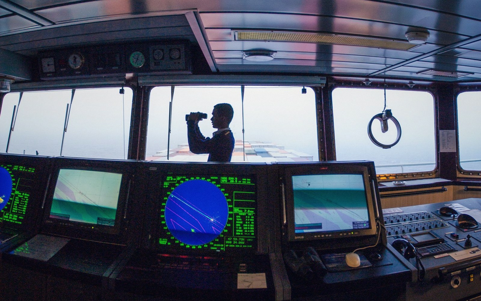 A ship's officer scans the horizon with binoculars from the bridge, surrounded by radar screens